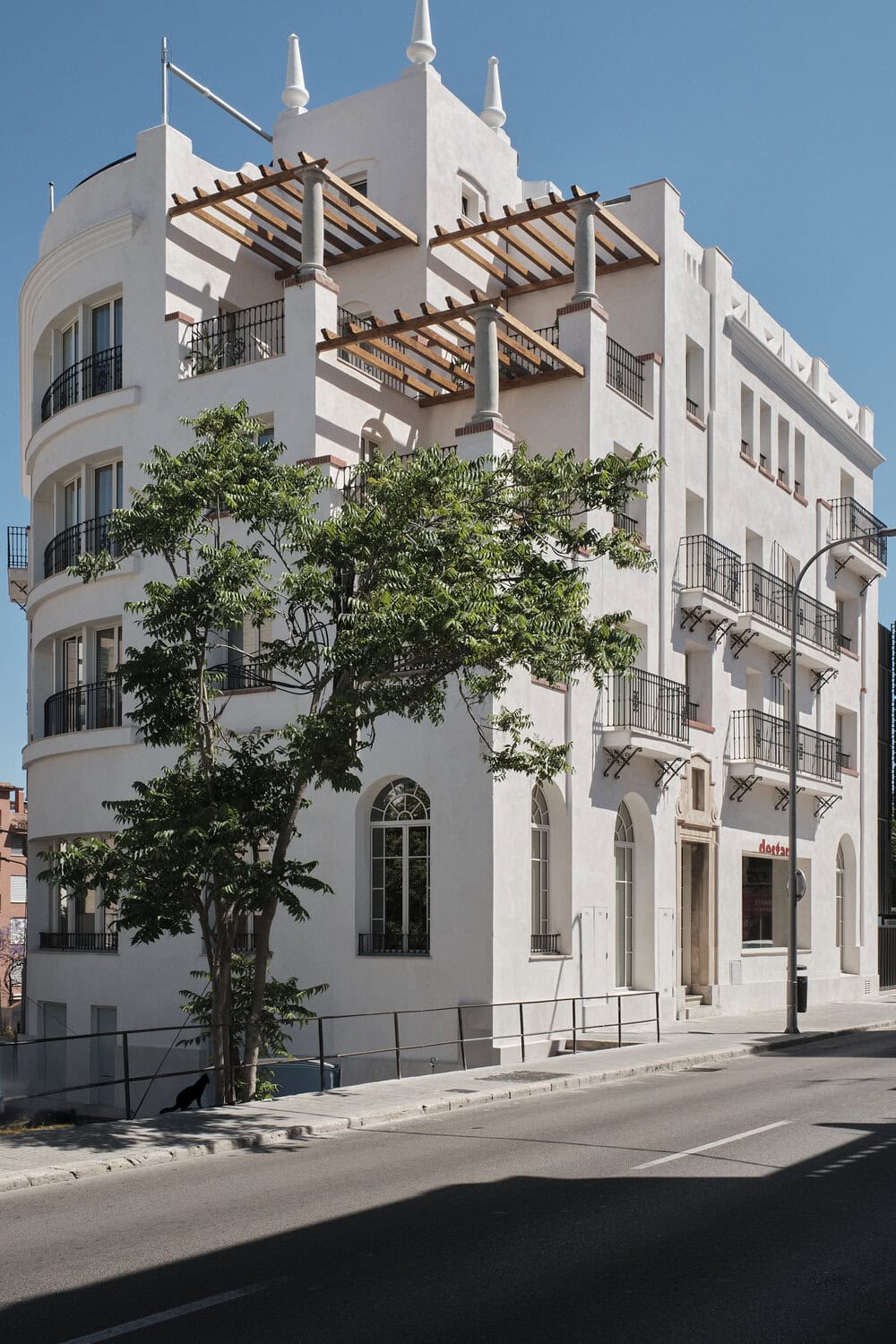 Weiß verputztes Eckgebäude im Art-déco-Stil mit Rundbalkonen, Dachpergola und Türmchen, davor Bäume an einer Straße bei blauem Himmel.