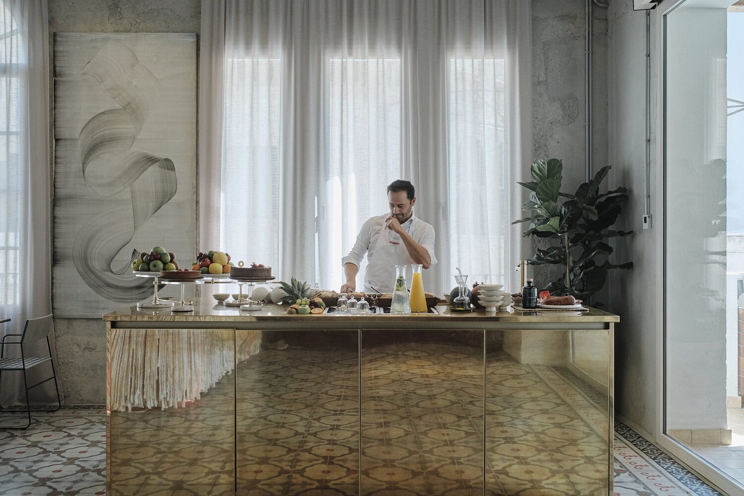 Koch bereitet an einer goldenen Theke vor bodenlangen Vorhängen ein Buffet mit Obst, Kuchen und Säften zu.