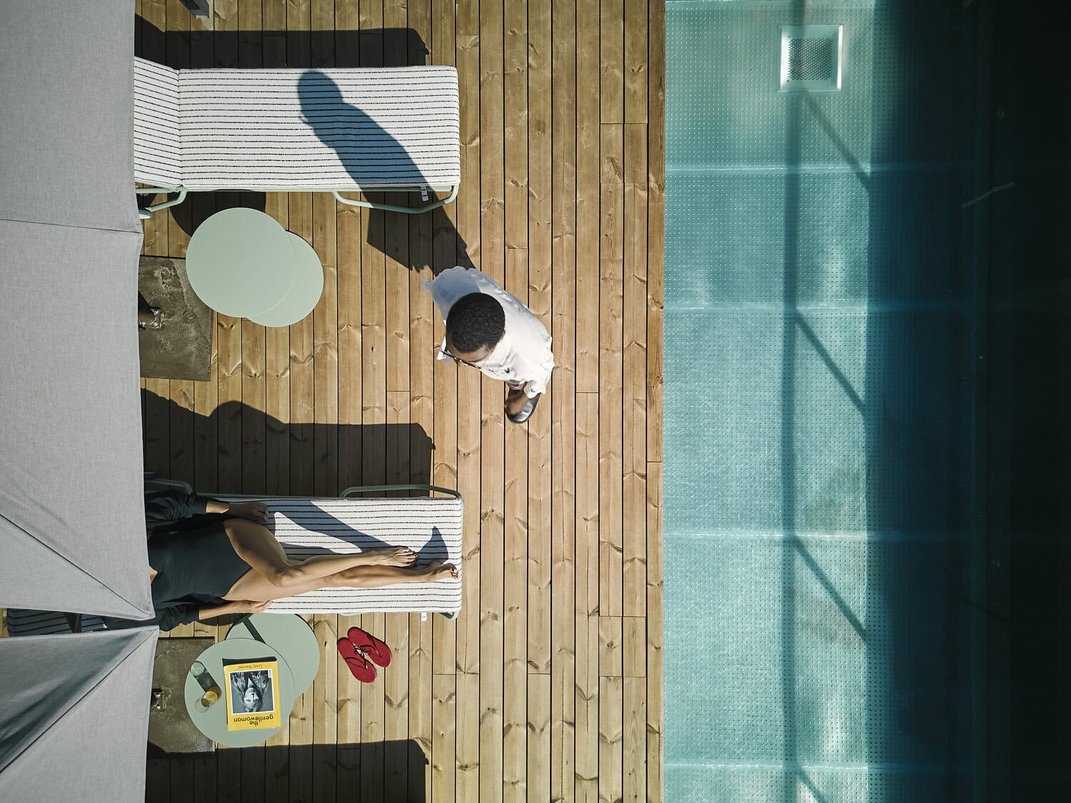 Draufsicht auf den Dachpool mit Holzdeck, gestreiften Liegen und zwei Personen, eine im Liegestuhl, eine stehend.