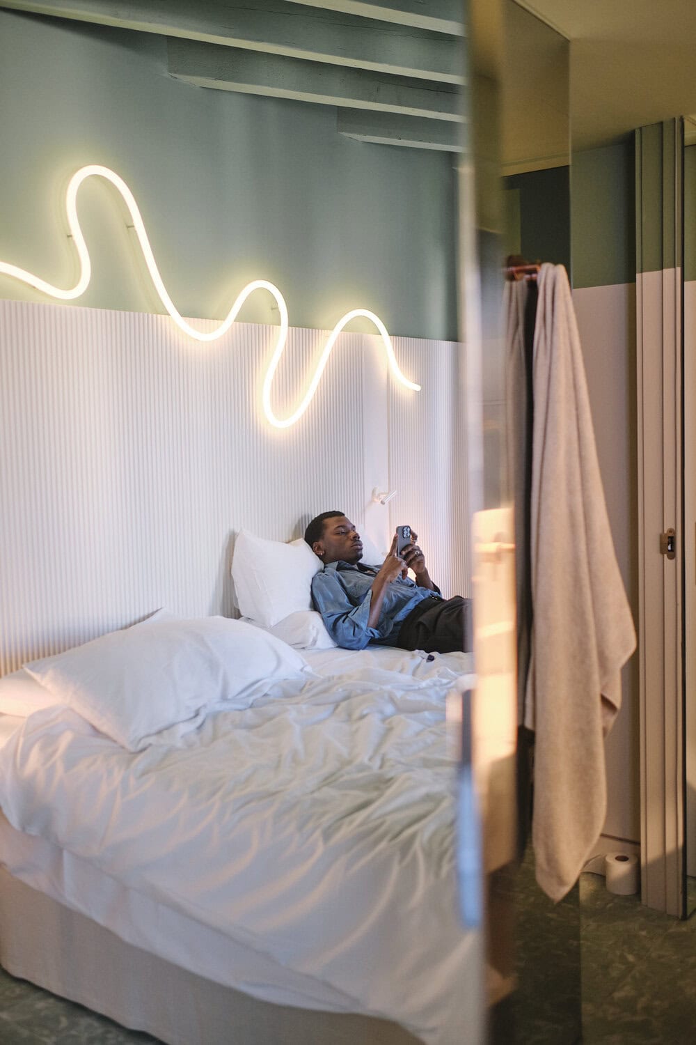 Mann liegt auf dem Bett eines Zimmers und schaut aufs Smartphone, über dem Bett eine geschwungene Neon-Leuchte.