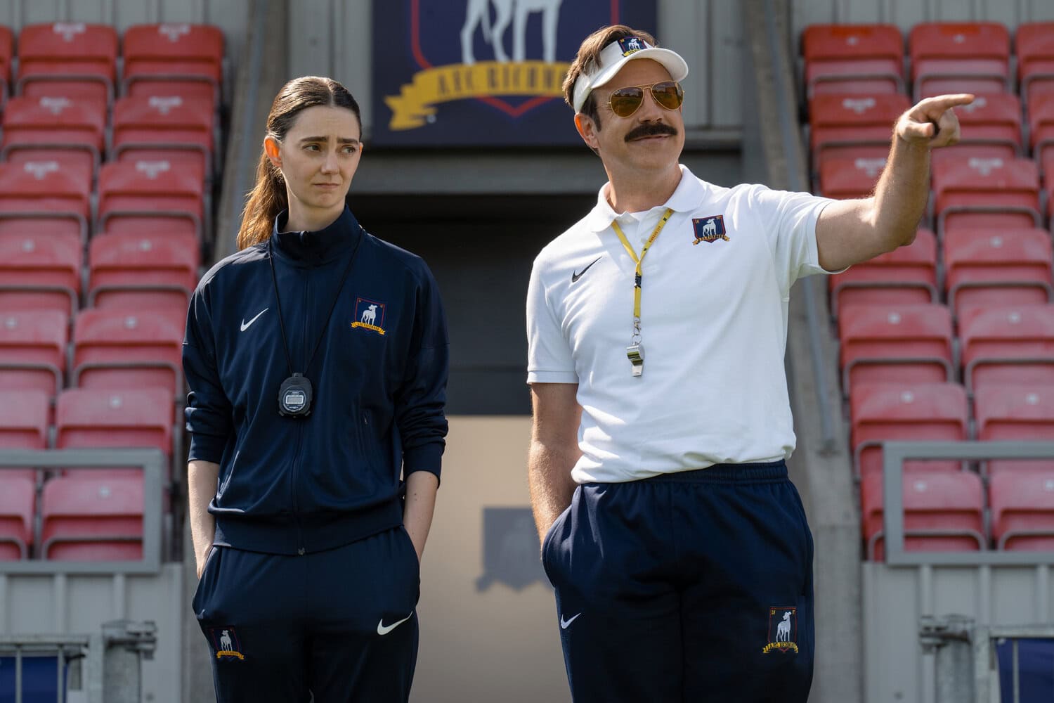 Szene aus Ted Lasso: Trainer mit Schirmmuetze zeigt auf etwas, neben ihm eine Frau im Trainingsanzug im leeren Stadion.