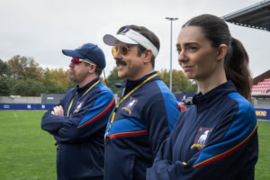 Szene aus Ted Lasso: Drei Trainer in blauen Trainingsjacken stehen nebeneinander am Spielfeldrand.