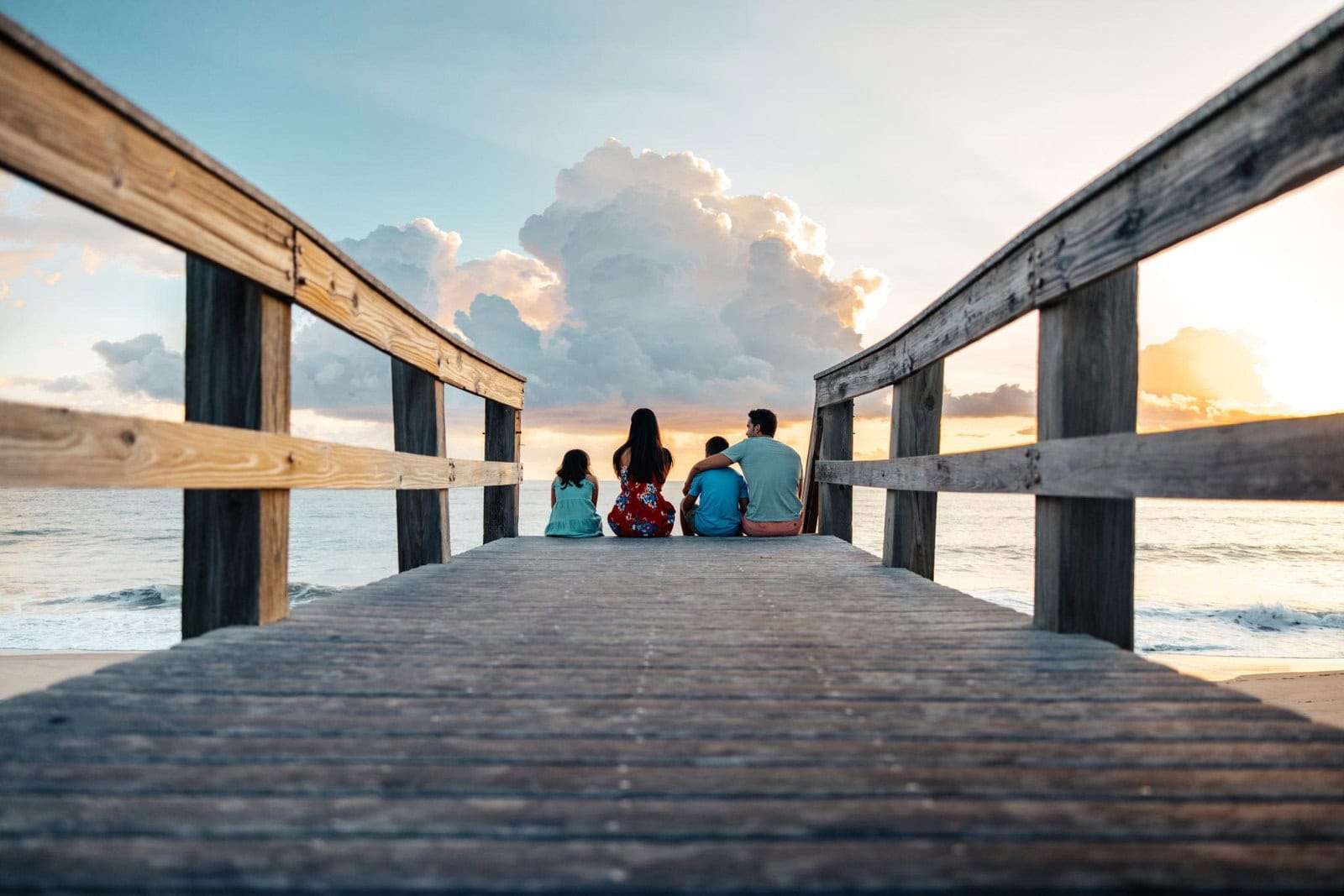 Familie sitzt von hinten gesehen am Ende eines Holzstegs am Vero Beach und blickt auf Meer und Wolken
