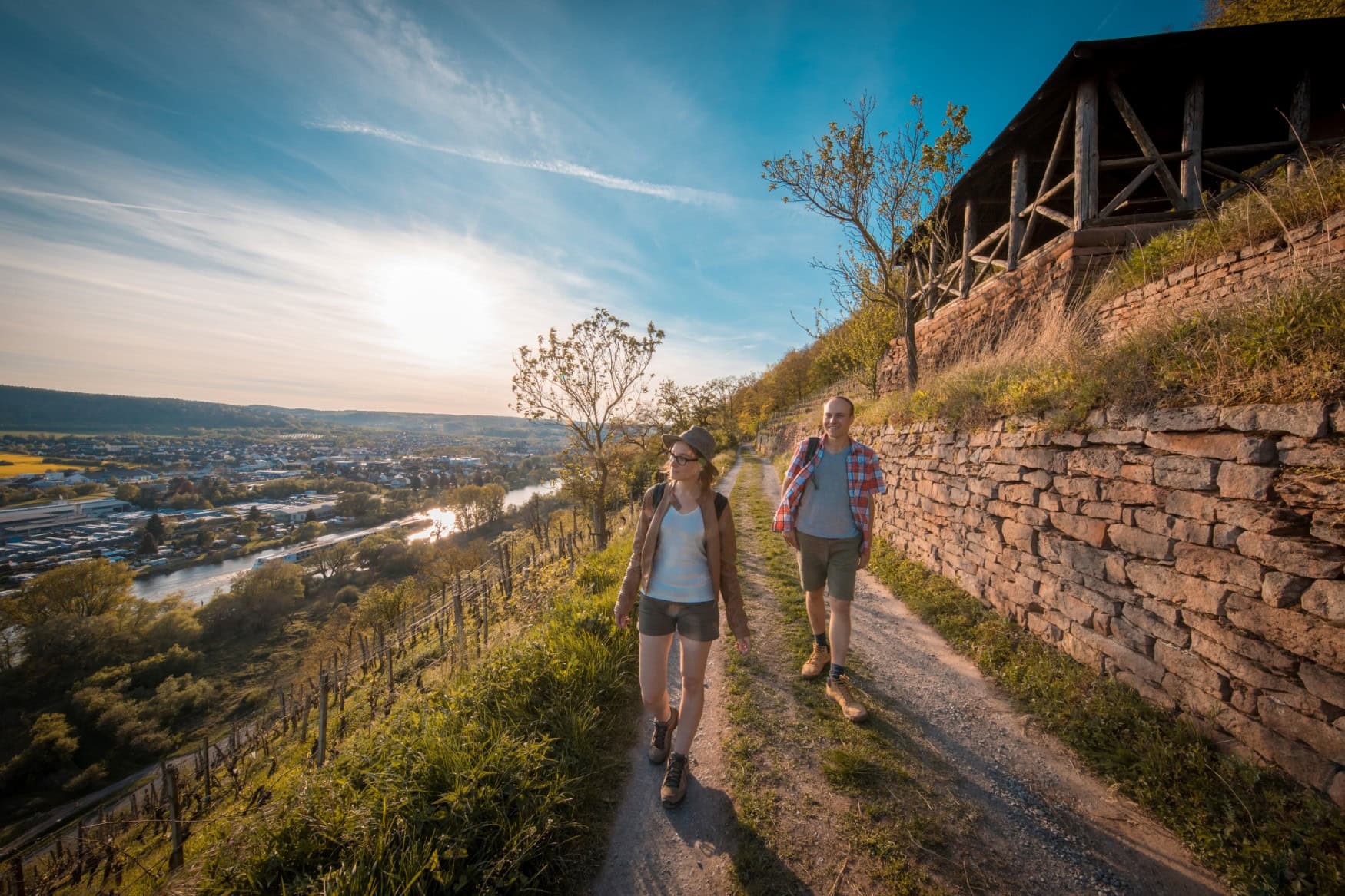 Zwei Menschen in sommerlicher Kleidung gehen auf einem Wanderweg Richtung der Kamera; rechts eine Mauer aus rotem Sandstein, links ein Hang mit Rebstöcken hinab ins Tal. Im Tal selbst fließt der main, dahinter eine kleine Ortschaft.