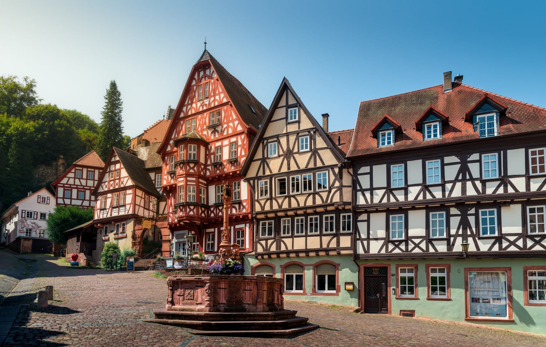 An einem Platz mit zentralem Brunnen stehen mehrere gut erhaltene Fachwerkhäuser in verschiedenen Farben nebeneinander, beschienen von der Sonne.