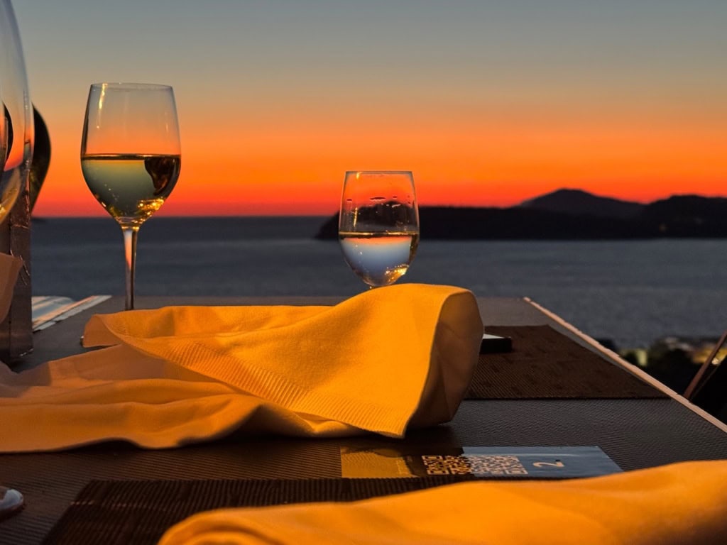 Nahaufnahme zweier Gläser Wein auf einem Tisch, dahinter unscharf die Adria im Sonnenuntergang.