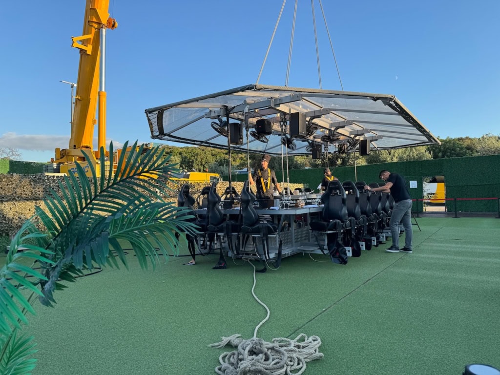 Ein langer Tisch mit Plexiglasdach wird gedackt. Das Dach ist mit Stangen am Tisch befestigt, am Dach wiederum hängen einige Tragseile, die zu einem Kran oberhalb des Bildrands führen.