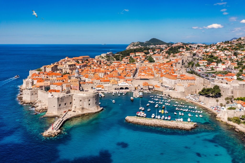 Luftaufnahme von Dubrovnik an einem sonnigen Tag, links die Adria, mittig die Stadt mit Stadtmauer und Hafenbecken