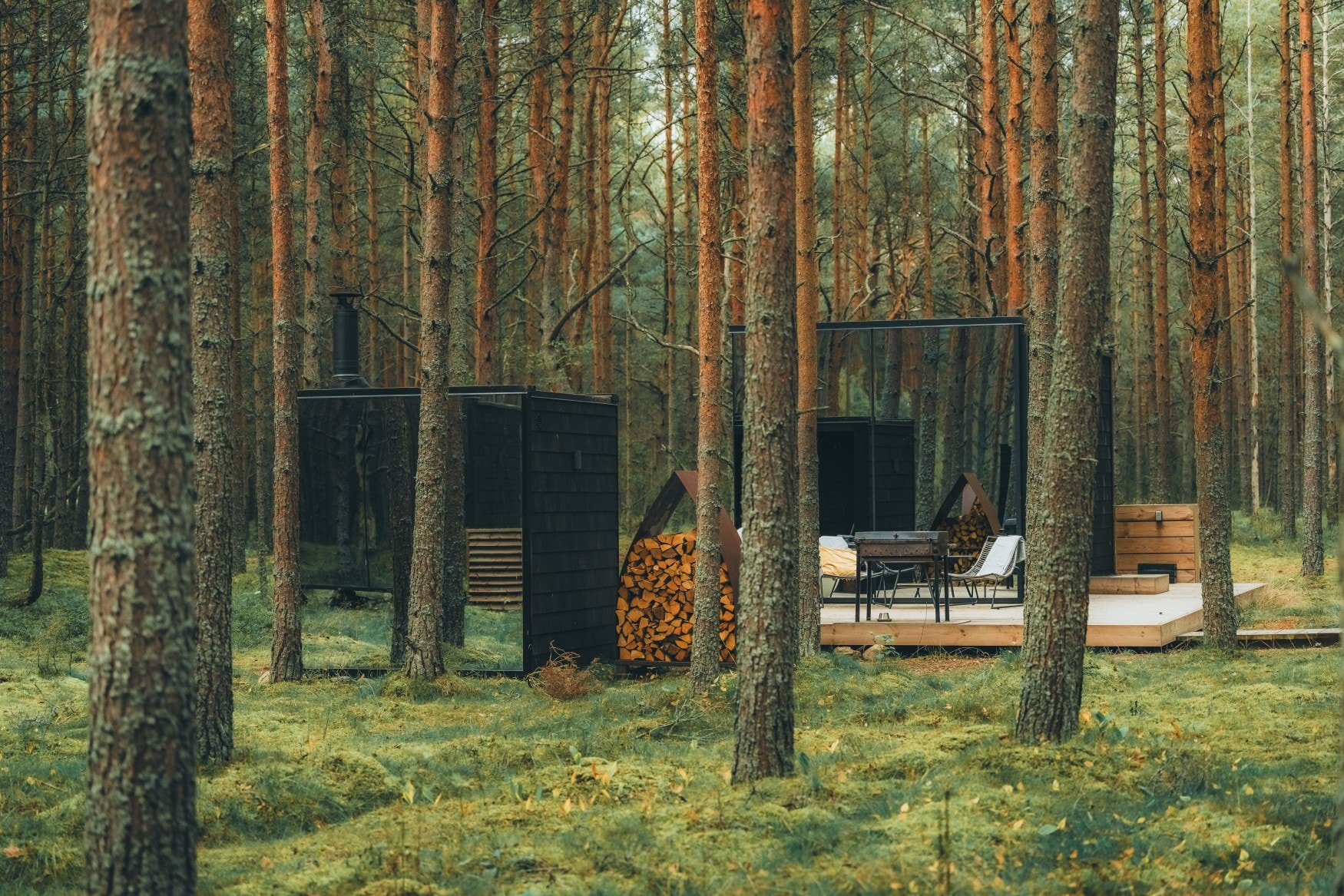 In einem grünen Wald steht eine verspiegelte Luxuslodge mit Holzverdeck und Grill.