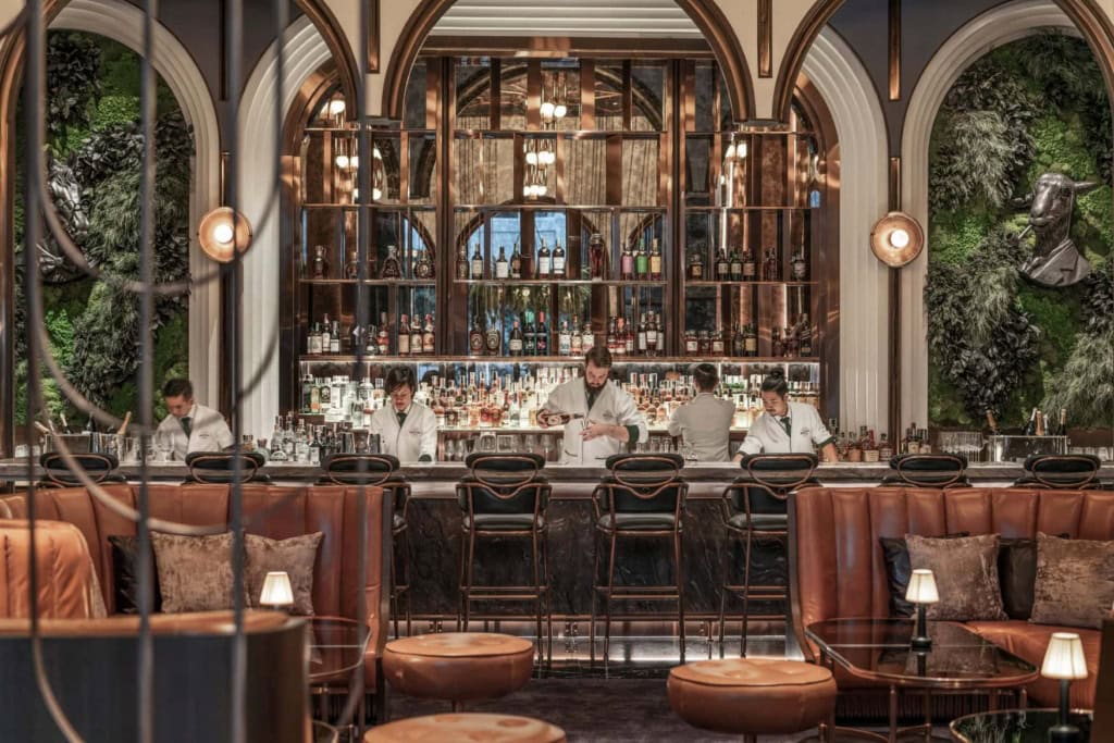 Hinter einer breiten, hohen Hotelbar mit Spiegelrückwand mischen mehrere Mitarbeiter in weißer Uniform Cocktails und Longdrinks.