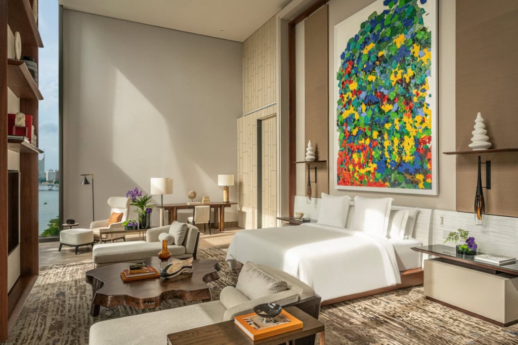 Blick in eine große, moderne Hotelsuite, mittig ein breites Bett, darüber ein gut zwei Meter hohes, modernes Gemälde. Vor dem Bett ein aus einem Baumstamm geschnittener Couchtisch, zu den Seiten je eine Recamiere.
