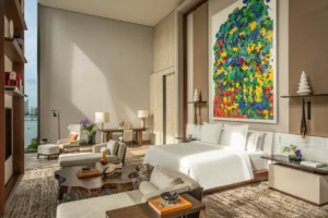 Blick in eine große, moderne Hotelsuite, mittig ein breites Bett, darüber ein gut zwei Meter hohes, modernes Gemälde. Vor dem Bett ein aus einem Baumstamm geschnittener Couchtisch, zu den Seiten je eine Recamiere.