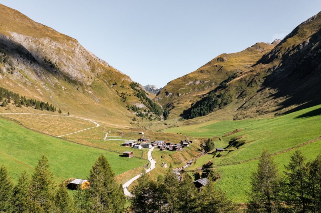 Die Fane Alm in Gitschberg Jochtal in Südtirol. Ein paar Häuser stehen in einem Tal neben einem kleinen Bach inmitten von Wiesen bedeckten Bergen.