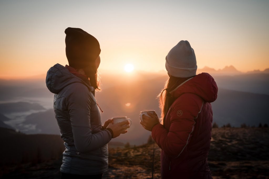 Zwei Personen mit Mützen stehen sich gegenüber und halten Tassen. Sie blicken auf die Vals und zwischen ihnen leuchtet die Sonne bei Sonnenaufgang durch.