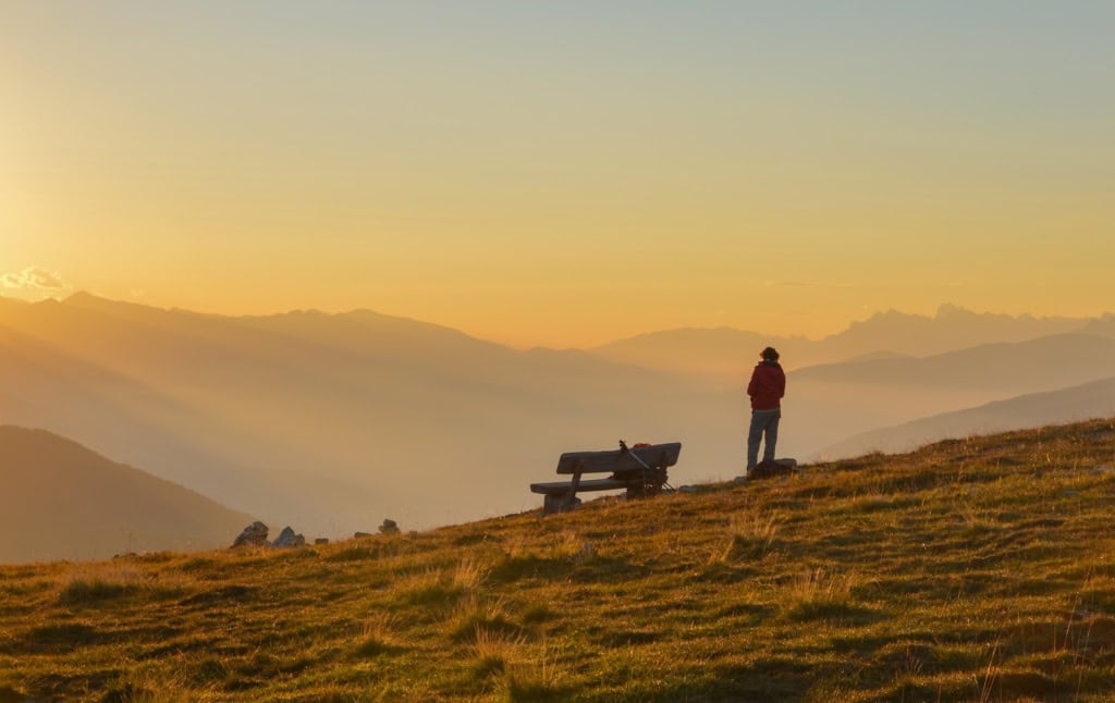 Eine wandernde Person macht Pause bei einer Bank, an die ihr Wanderstock anlehnt, und schaut beim Sonnenaufgang in Vals auf die Silhouetten der Alpen in Gitschberg, Jochtal