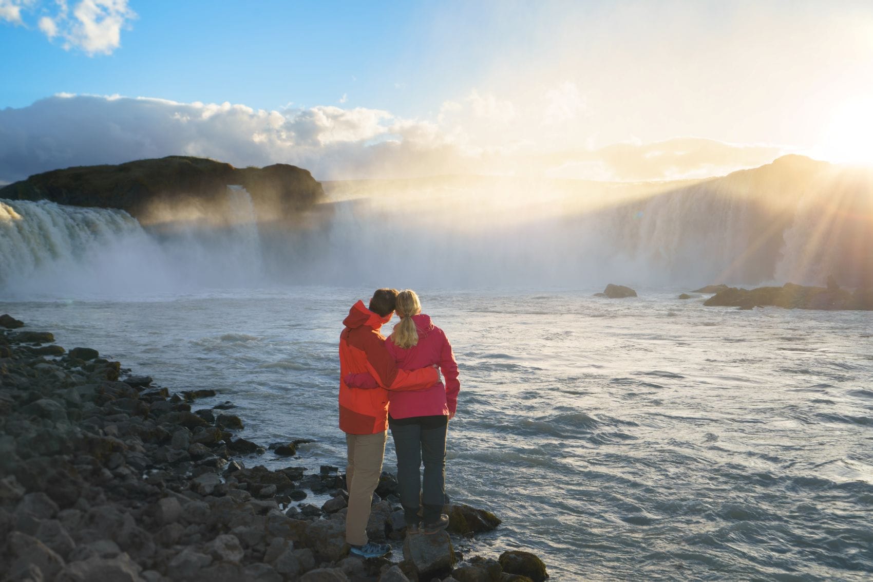 Zwei Besucher genießen den Blick auf den beeindruckenden Goðafoss-Wasserfall in Nordisland bei tief stehender Sonne.