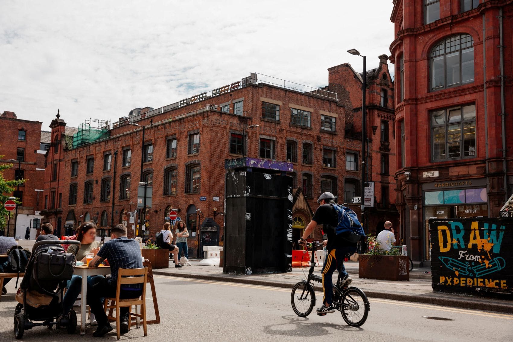 Straßenszene im Northern Quarter in Manchester mit Cafés, Radfahrer und historischer Backsteinarchitektur.