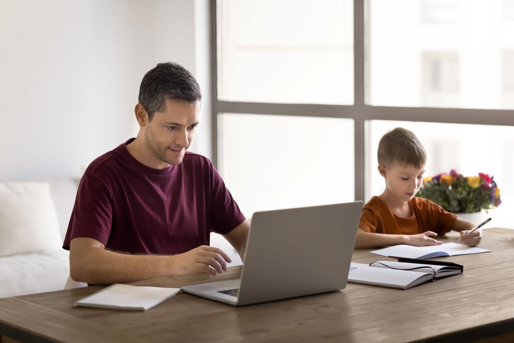 Vater arbeitet an einem Laptop im Homeoffice, während sein Sohn am selben Tisch Hausaufgaben macht.