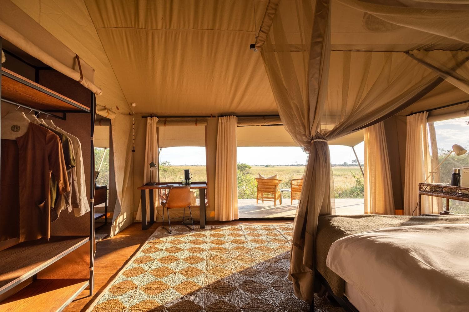 Innenraum eines Safari-Zelts mit Himmelbett, Garderobe und Schreibtisch unter cremefarbenem Stoffdach, durch geöffnete Türen Blick in die weite Grassavanne im Abendlicht.