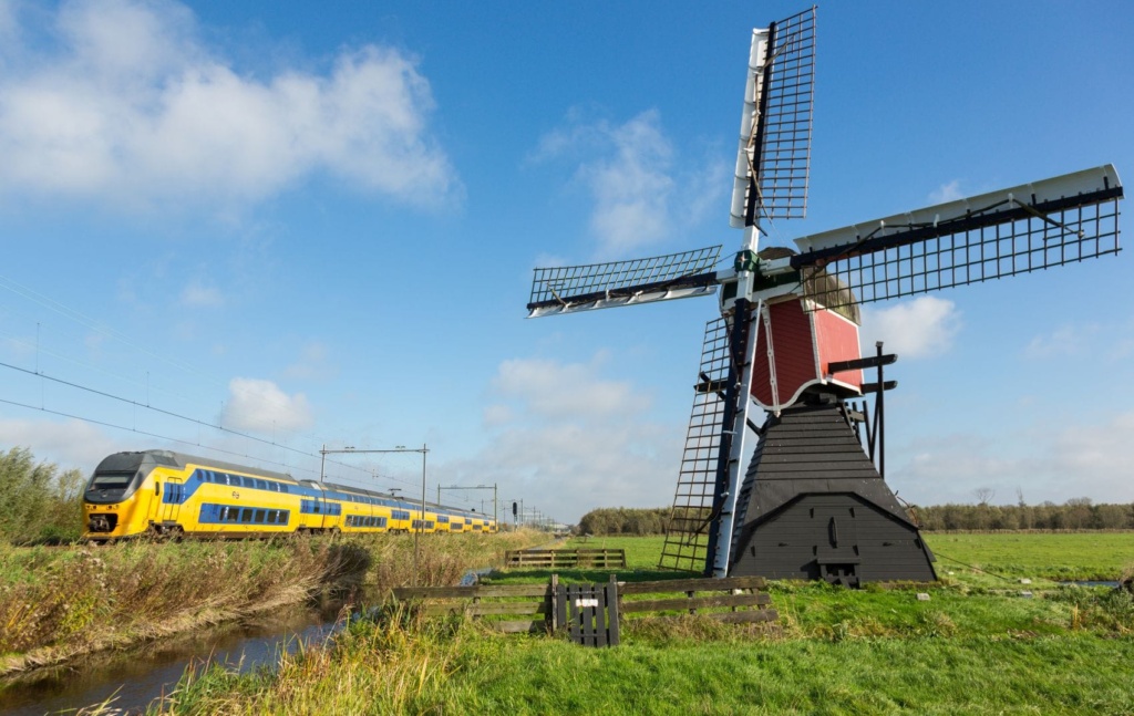 Ein gelb-blauer Zug der niederländischen Bahngesellschaft NS fährt an einer historischen Windmühle durch die niederländische Landschaft.