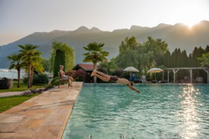 Ein Mann springt kopfüber vom Rand eines Hotelpools ins Wasser. Am Beckenrand sitzt eine Frau und schaut ihm zu. Im Hintergrund sind die Alpen sichtbar.