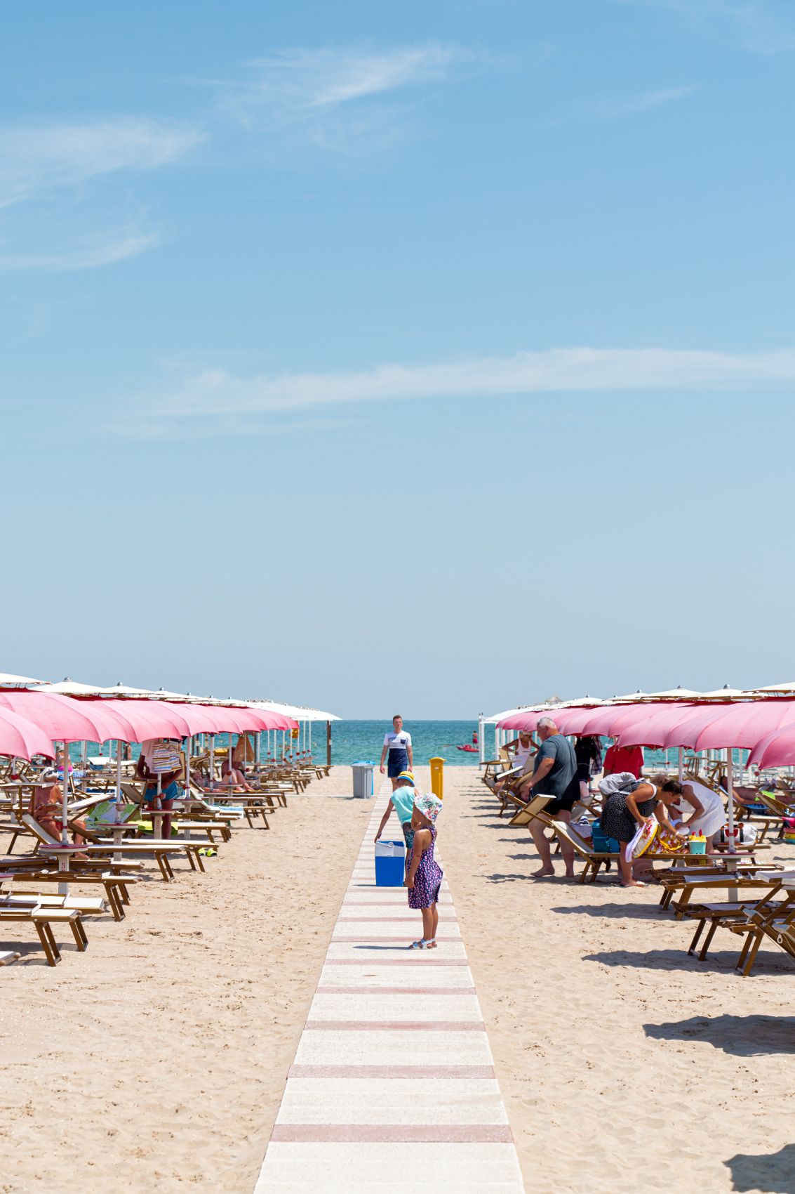 Familien und Badegäste am breiten Sandstrand von Rimini an der Adriaküste. Rosafarbene Sonnenschirme säumen den Weg zum Meer.