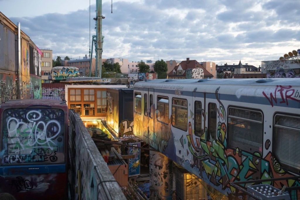 Zwei mit Graffiti besprühte U-Bahn-Waggons stehen im Abendlicht auf dem Gelände des Schlachthofviertels in München, im Hintergrund Wohnhäuser und ein beleuchtetes Gebäude.