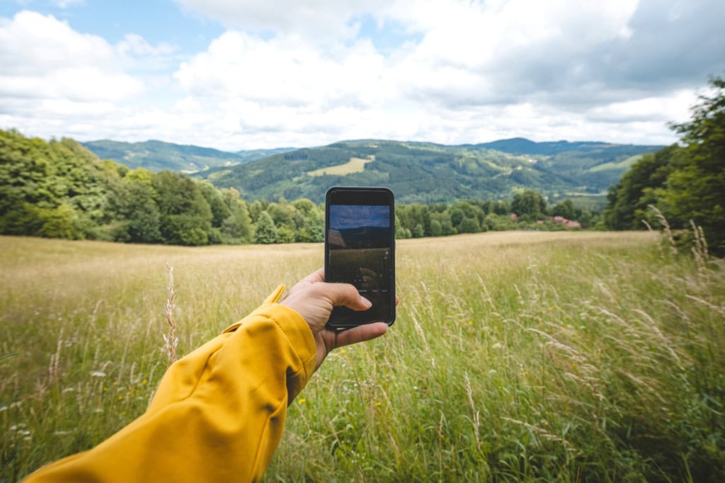 Eine Hügellandschaft mit gelben Feldern und grünen Wäldern. Eine Person hält ein Smartphone vor die Kamera und nimmt ein Foto auf.