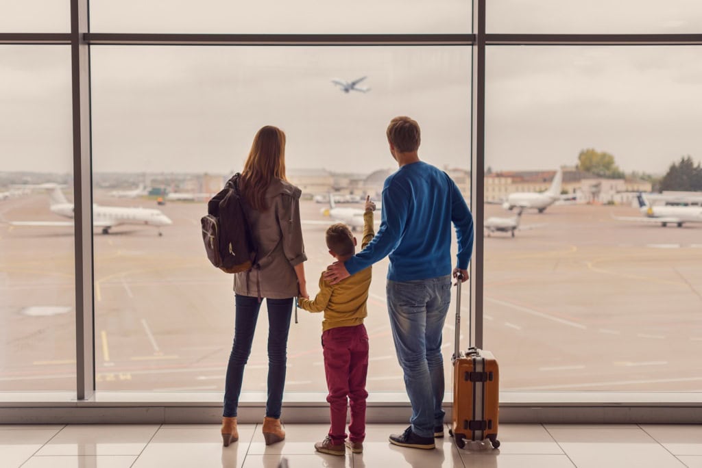 Ein Mann, eine Frau und ein Kind im Grundschulalter stehen mit ihrem Handgepäck an einer großen Scheibe enes Flughafens und betrachten die abhebenden Flugzeuge.