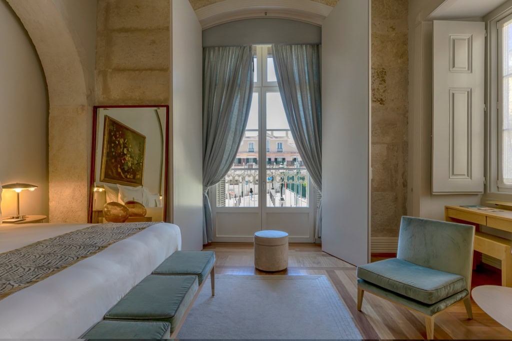 Ausschnitt aus einem Hotelzimmer im Verride Palácio in Lissabon; zentral ein hohes Doppelfenster, links ein freistehendes Bett, rechts ein flacher, blauer Polstersessel.