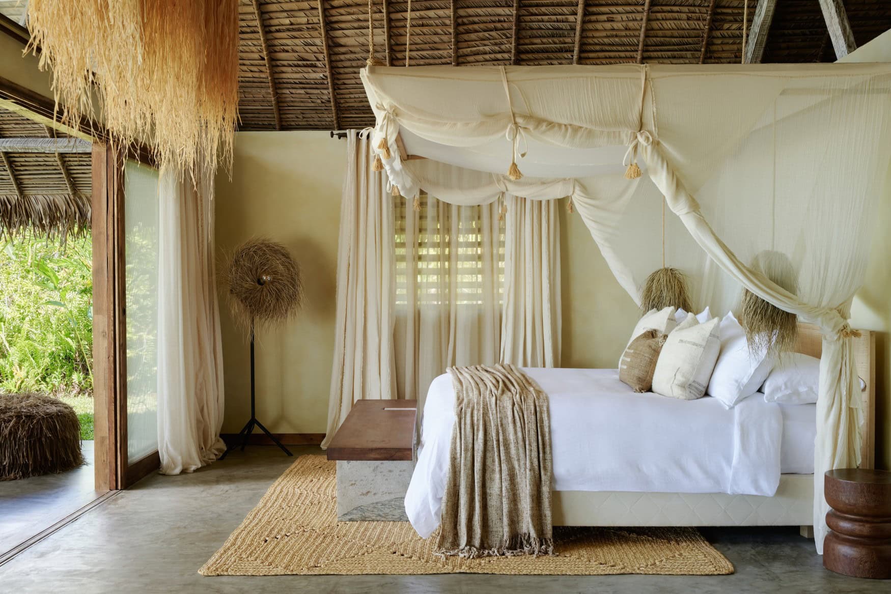 Schlafzimmer eines Bungalows im Voaara, Madagaskar, mit großem Himmelbett unter Strohdach, weißen Leinenbaldachinen, natürlichen Textilien in Beige und Blick in den grünen Außenbereich.