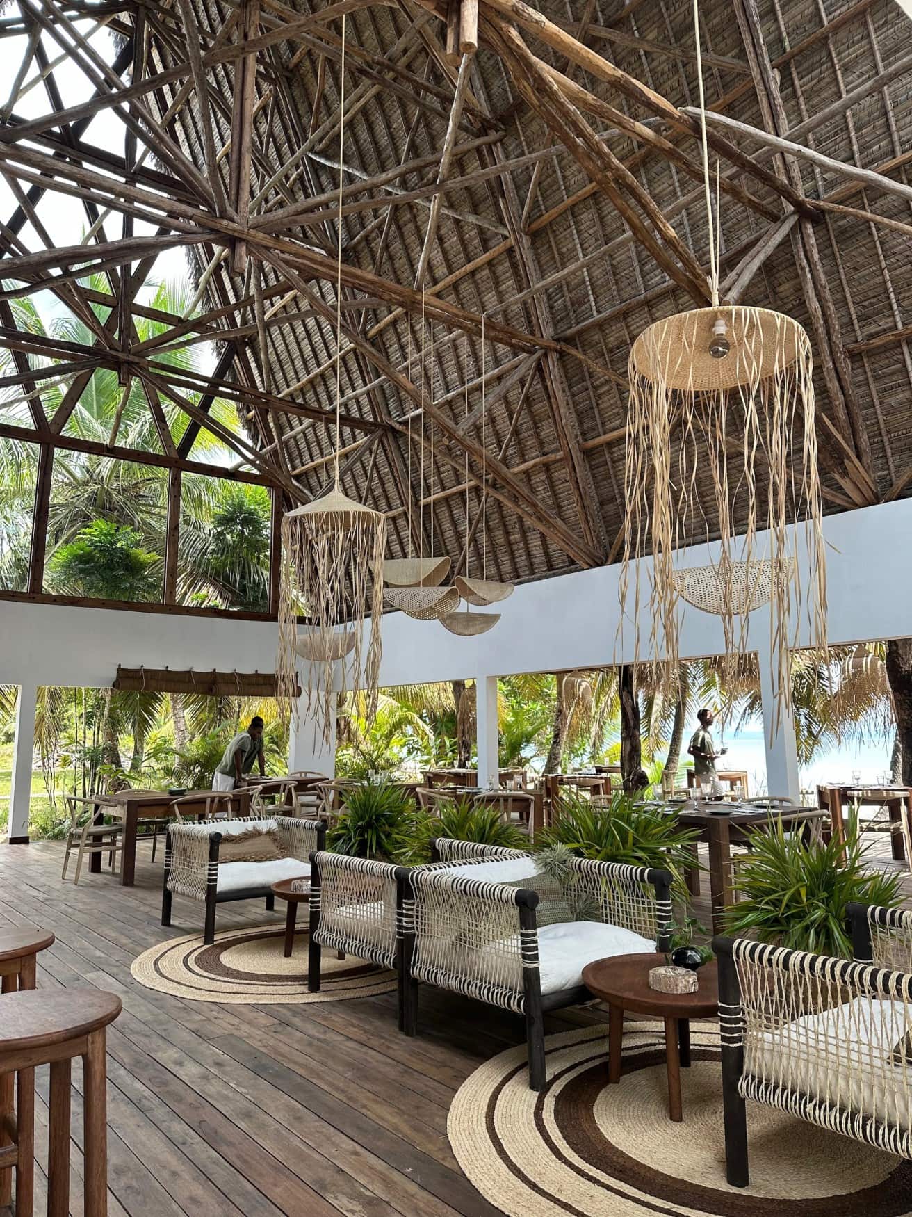 Innenbereich des Restaurants im Voaara auf Madagaskar mit hohem offenem Strohdach aus Naturholzkonstruktion, hängenden Rattanleuchten, geflochtenen Sesseln auf Holzdeck und Meeresblick durch die offenen Seiten.