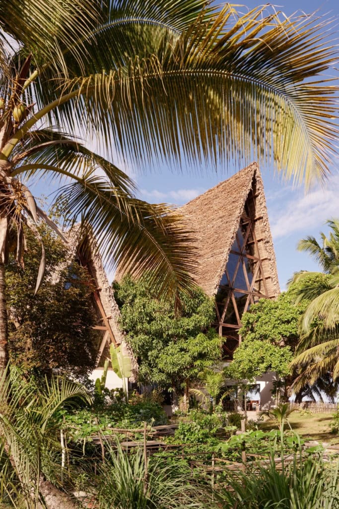 Außenansicht des charakteristischen spitzen Strohdachs der Villa im Voaara, Madagaskar, eingebettet zwischen Palmen und Tropengarten mit kleinem Gemüsegarten im Vordergrund.