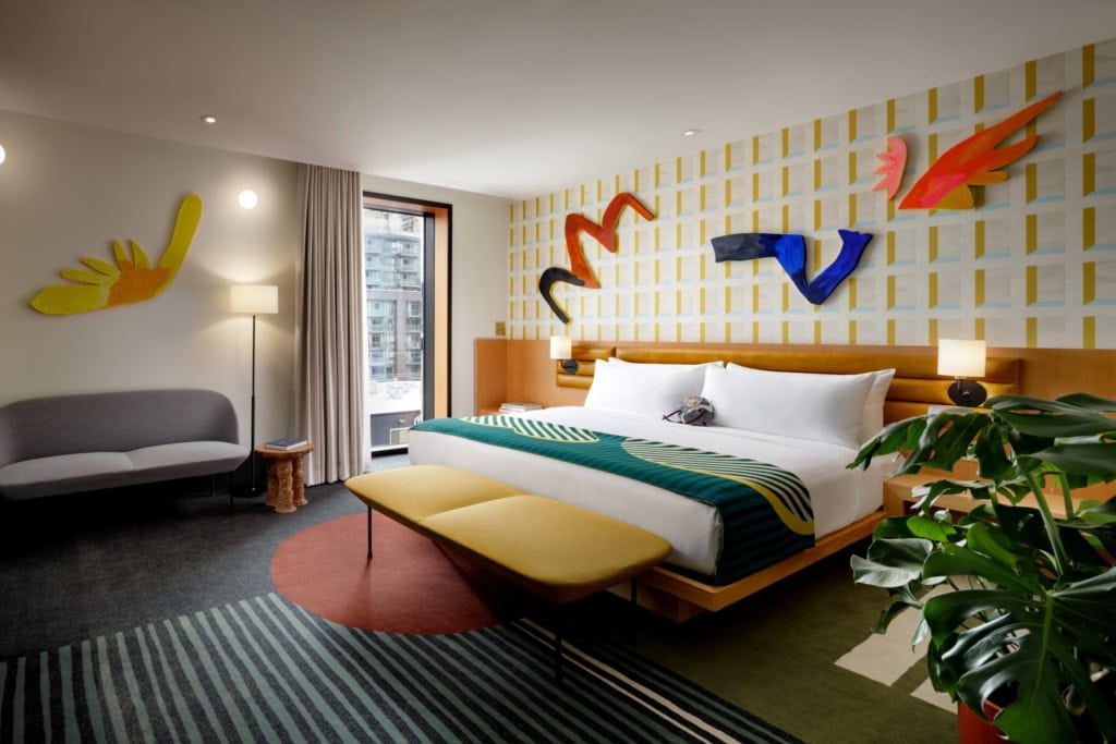 Suite im Hotel The Drake in Toronto; die Wand hinter dem breiten Bett ist mit einem geometrischen Muster verziert, darauf einige bunte Kunstelemente; auch der Teppichboden trägt ein auffälliges Farbmuster.